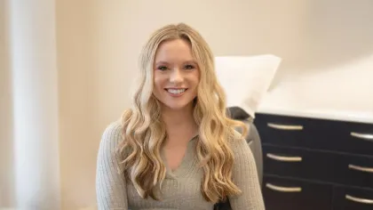 Smiling young woman with long blonde hair sitting in a modern office chair in a bright room.